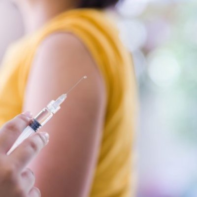 Close-up of vaccine with women in background with yellow shirt sleeves rolled up.